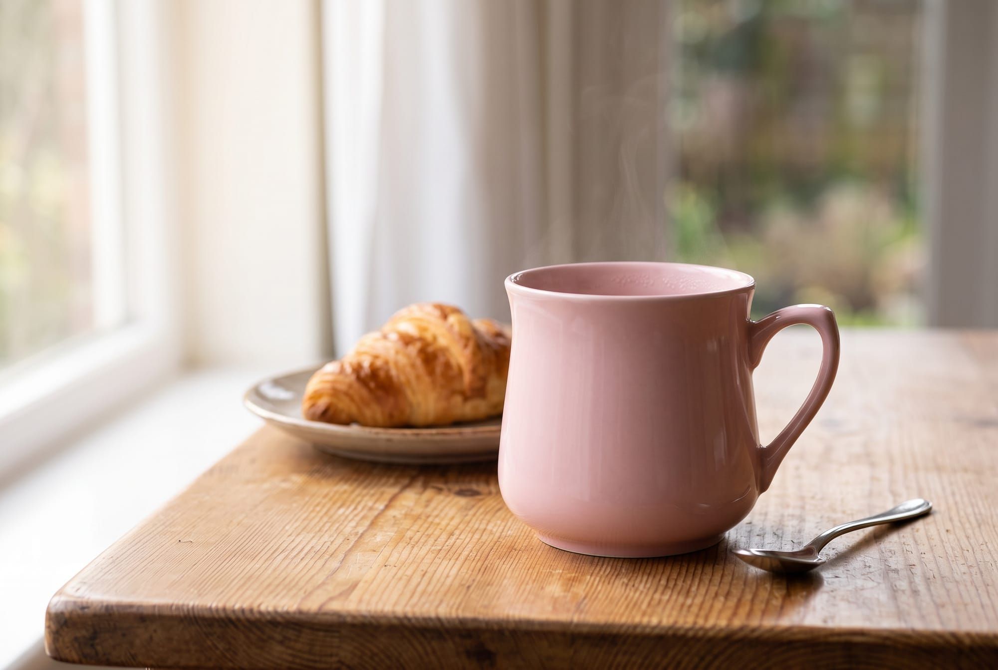 The same pink mug on a worn wooden table by a window, with a croissant on a plate behind it and a spoon beside it